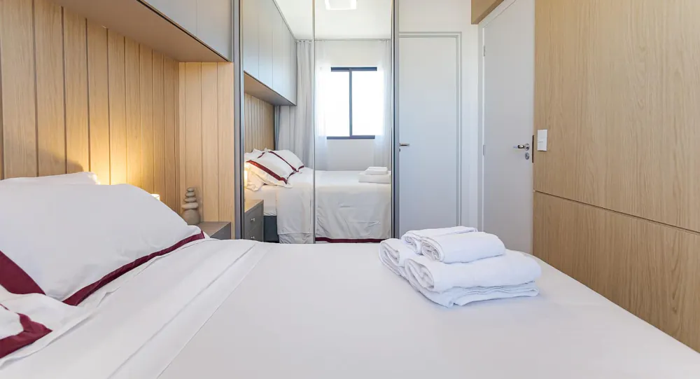 Detalhe do enxoval: lençol branco com faixa bordô e toalhas dobradas sobre a cama