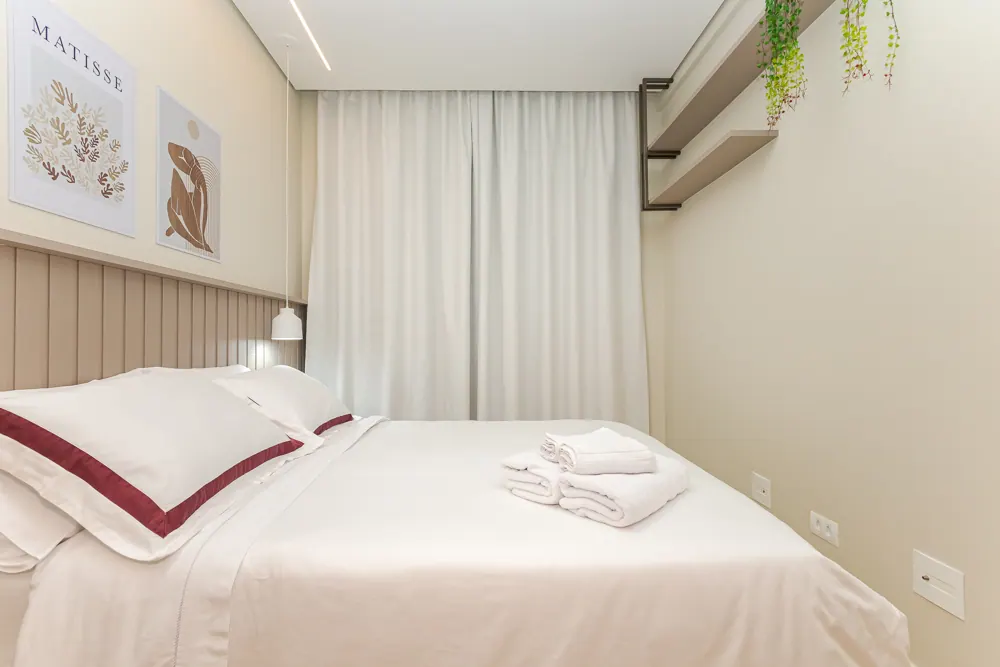 Toalhas brancas dobradas sobre a cama arrumada, fronhas com faixa bordô e quadros de Matisse