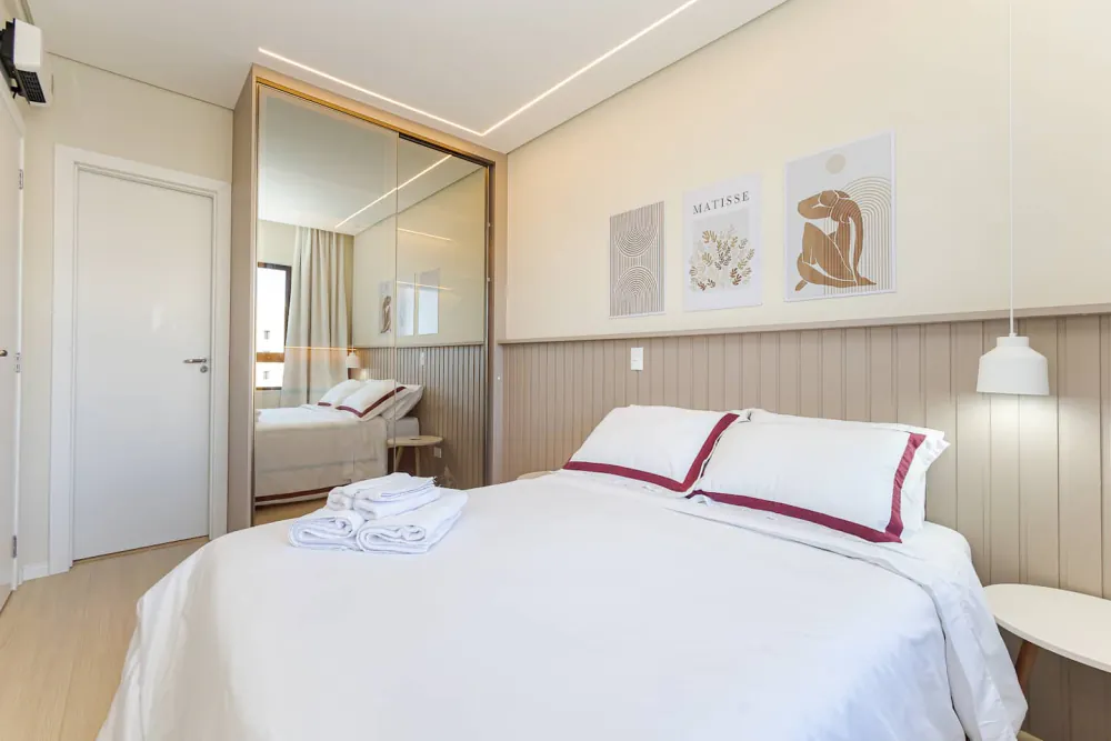 Quarto de um dos apartamentos da Rhose: cama arrumada com enxoval branco de faixa bordô e toalhas dobradas