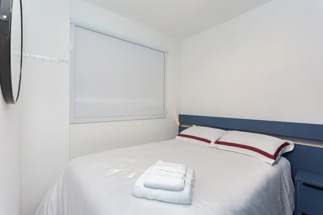 Cama de casal com enxoval branco e faixa bordô, toalhas dobradas e cabeceira azul