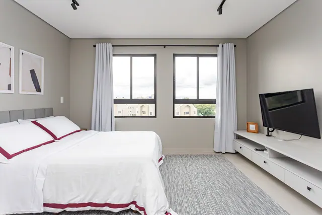 Quarto amplo com cama de enxoval branco e faixa bordô e janelas com vista
